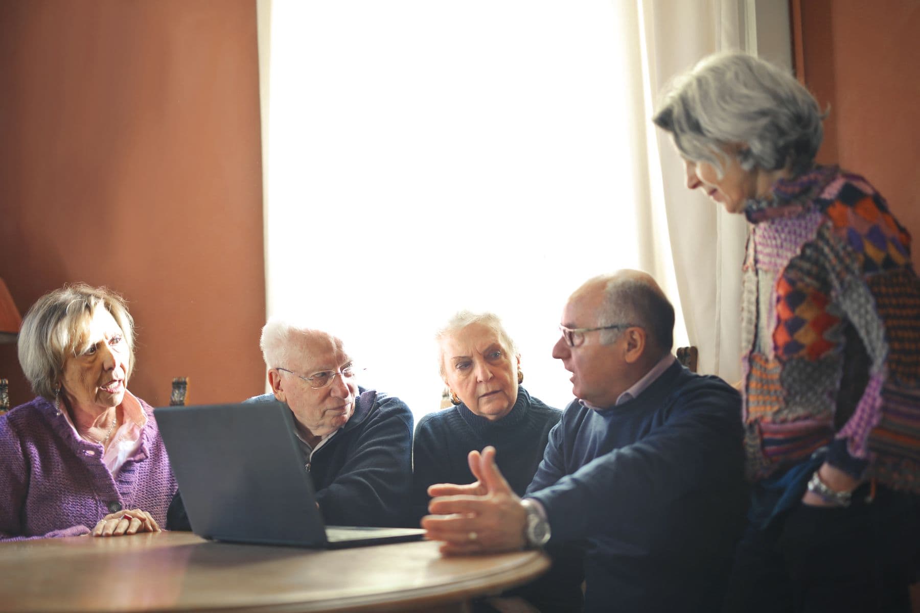 Five older people around a table looking at the same laptop, one of them mid-sentence