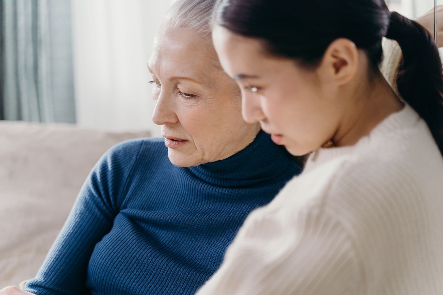 An older woman and a younger woman sitting close together on a sofa, looking down